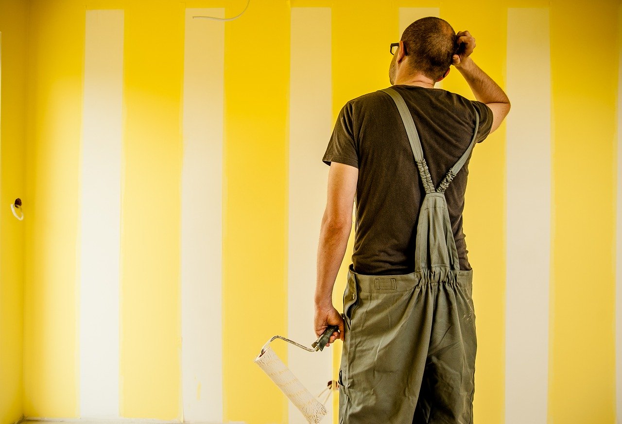 Hombre con un rodillo en la mano, mirando una pared parcialmente pintada y rascándose la cabeza